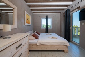 A bedroom with a wooden bed, a bedside table with a lamp, a window with a view, and a balcony door.