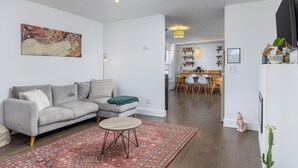 Een moderne woonkamer met een grijze bank, een ronde salontafel en een tapijt met een rood patroon. Er is een eethoek met een houten tafel en stoelen, en een keuken is zichtbaar op de achtergrond.