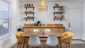 Een eetruimte met een houten tafel, vier stoelen, een koffiezetapparaat en planken met keukengerei en boeken.