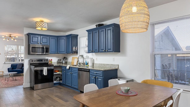 Een moderne keuken met blauwe kastjes, roestvrijstalen apparatuur en een eethoek met een houten tafel en gele stoelen.