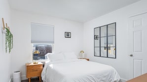 Een minimalistische slaapkamer met een bed, nachtkastjes, een raam met zonwering, een spiegel en een deur.