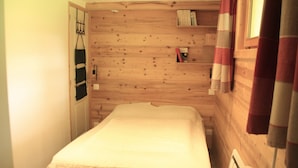Ein kleiner, gemütlicher Raum mit einer Holzwand, einem Bett mit weißer Bettdecke, einem Regal mit Büchern und einem Fenster mit Vorhängen.
