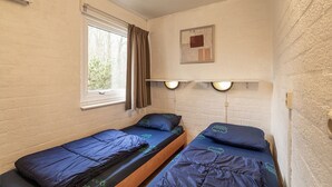 Una habitación pequeña y acogedora con dos camas individuales, una ventana con cortinas, un estante en la pared con un cuadro enmarcado y una pared de ladrillo blanco.