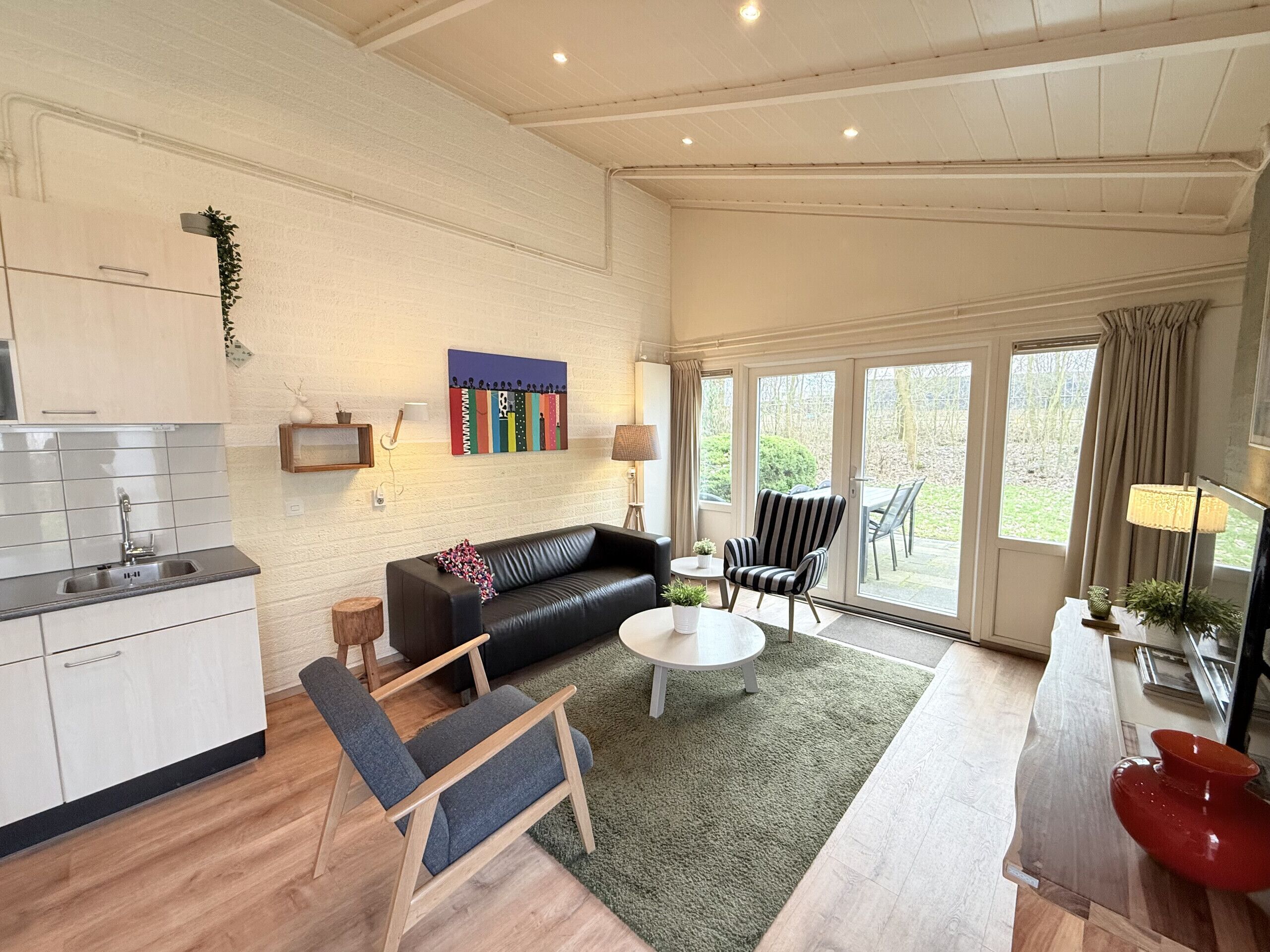 Una sala de estar moderna con un sofá de cuero negro, una mesa de centro de madera y un sillón a rayas. Se ve una cocina a la izquierda, con armarios blancos y un salpicadero de azulejos.