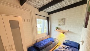 A compact room with two beds, a window, a radiator, and a shelf with a plant.