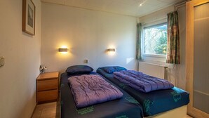 A bedroom with two beds, a wooden nightstand, a wall-mounted light, and a window with curtains. - Bungalowpark Simpelveld - Schakel 5 (Simpelveld)