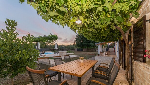 Une terrasse extérieure avec une table et des chaises en bois, entourée d’un sol en pierre et de verdure.