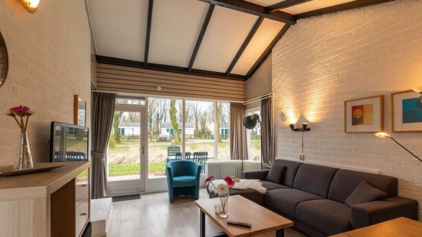 A modern living room with a sofa, a coffee table, and a TV. There is a window with curtains, a blue chair, and a lamp. - Bungalowpark Simpelveld - Castra 47 (Simpelveld)
