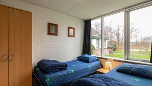 Une chambre avec deux lits, une armoire en bois, une fenêtre donnant sur l’extérieur et une petite table avec une lampe.