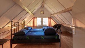 Un lit simple dans une chambre mansardée au plafond haut, avec vue sur l’extérieur par une fenêtre.