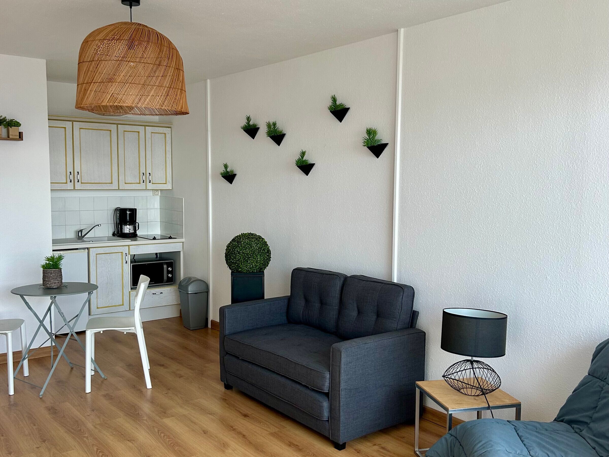A modern living room with a grey sofa, a small round table, and a lamp. There is a kitchen area with white cabinets and a coffee maker.