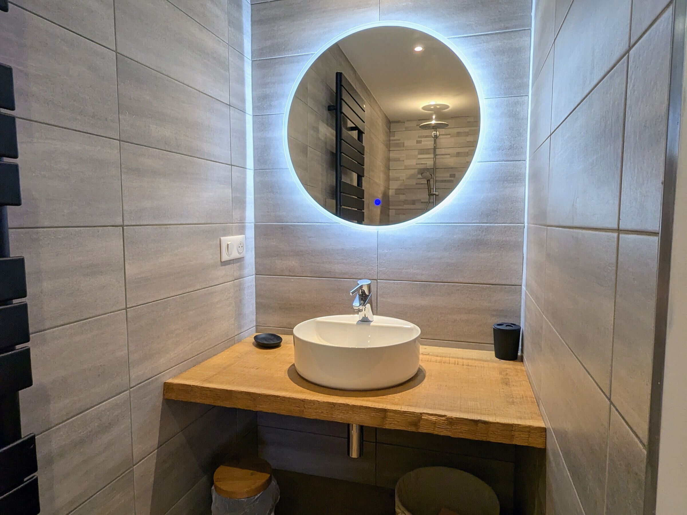Une salle de bain moderne avec un miroir rond, un lavabo blanc et un plan de toilette en bois.