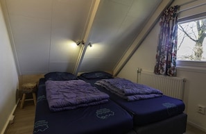 A small, single-room accommodation with two beds, a window with curtains, a radiator, and a small stool. - Bungalowpark Simpelveld - Alpha 64 (Simpelveld)