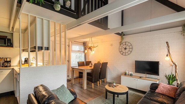 Una sala de estar moderna con sofá, mesa de centro y televisor. Hay un comedor con mesa y sillas. La habitación tiene una escalera y una zona de cocina con refrigerador y microondas.