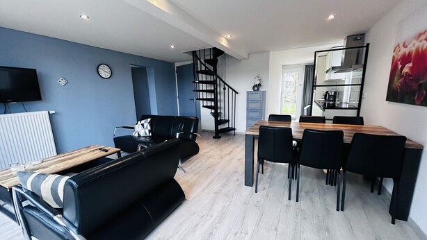 A modern living space with a black leather sofa, a wooden dining table with chairs, a staircase, a kitchen area, and a television mounted on the wall. - Bungalowpark Simpelveld - Delta 125 (Simpelveld)