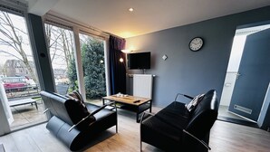 A modern living room with two black leather sofas, a wooden coffee table, a TV on the wall, and a large window offering a view of the outdoors. - Bungalowpark Simpelveld - Delta 125 (Simpelveld)