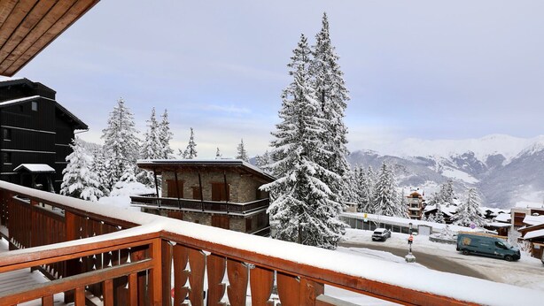 Ein Holzbalkon mit schneebedeckten Geländern mit Blick auf einen Bergort mit schneebedeckten Bäumen und Gebäuden.