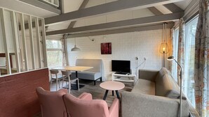 A modern living room with a high ceiling, exposed beams, a grey sofa, a pink chair, a small round table, a TV on a white stand, and a window with curtains. - Bungalowpark Simpelveld - Alpha 77 (Simpelveld)