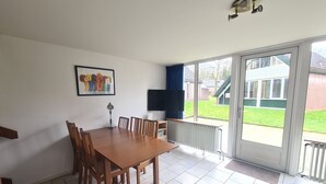 A dining area with a table and chairs, a TV, and a large window with a view of a house and lawn. - Bungalowpark Simpelveld - Delta 109 (Simpelveld)