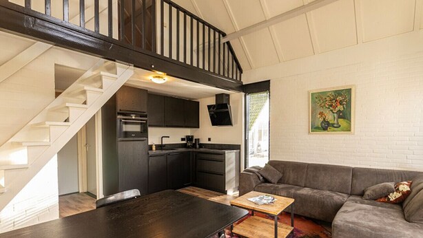 Een modern interieur met een trap naar een slaapkamer op een entresol, een keukenhoek en een woonkamer met een bank en salontafel.