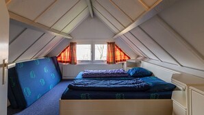 Een kleine, driekante slaapkamer op zolder, met een bed, een stoel en een raam met gordijnen.