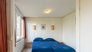 A small, single-room hotel room with a bed, a window with curtains, a wall-mounted light, and a small table. - Bungalowpark Simpelveld - Delta 148 (Simpelveld)