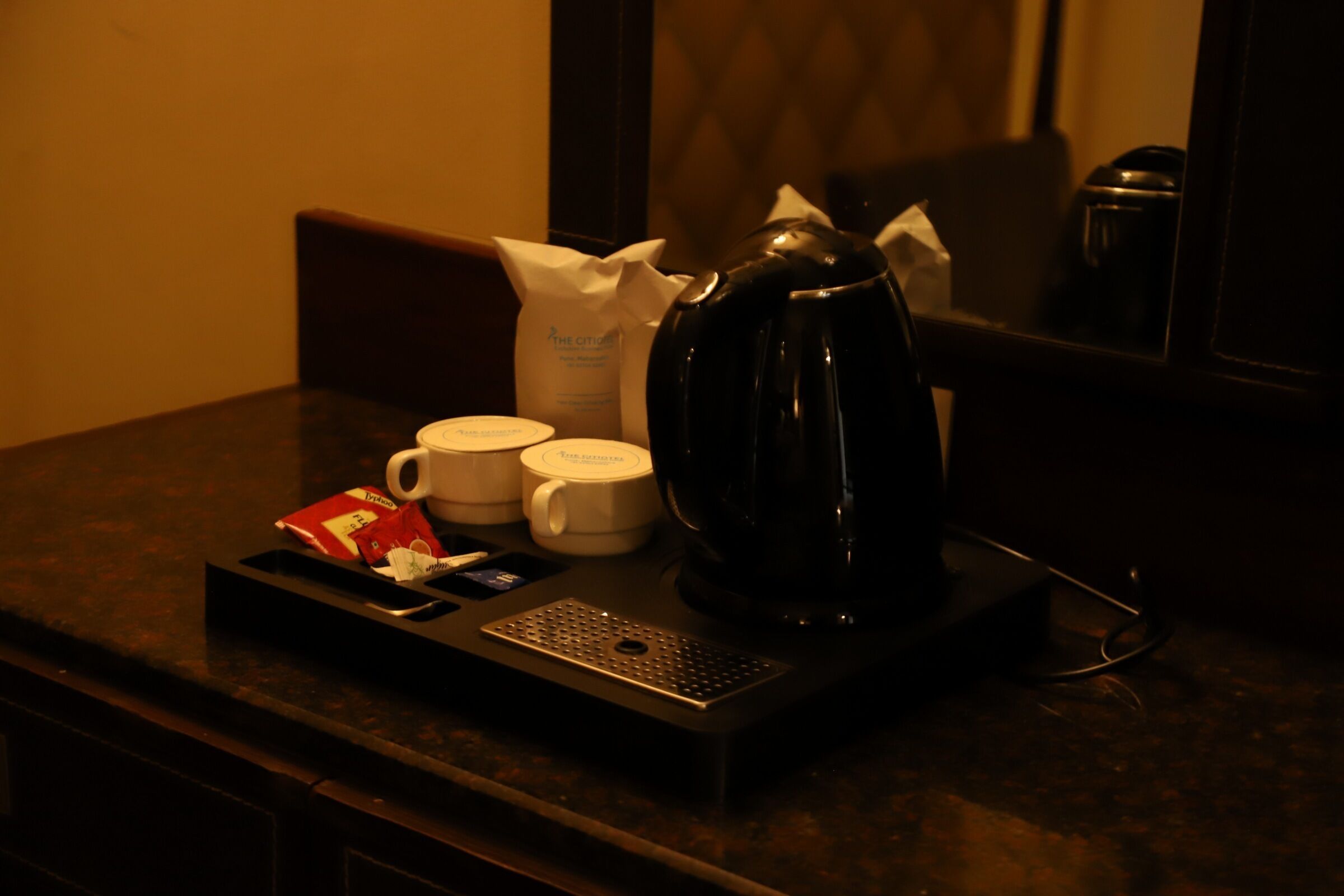 A coffee maker, two cups, a tray with a packet, and a bag on a dark countertop.