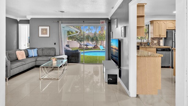 A modern living room with a grey sofa, a glass coffee table, and a TV. There is a glass door leading to a pool area with a hammock and palm trees. The kitchen has wooden cabinets and stainless steel appliances.