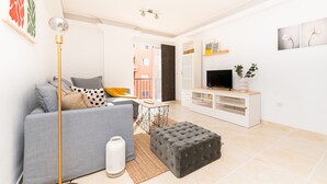 Una sala moderna con un sofá gris, un otoman negro, una lámpara de pie, un televisor sobre un mueble blanco y una pequeña mesa con una planta.