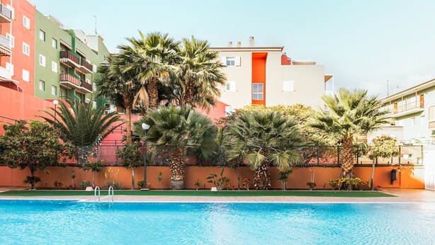 Una piscina con superficie de agua azul cristalina, rodeada de palmeras y un edificio de varios pisos con balcones.