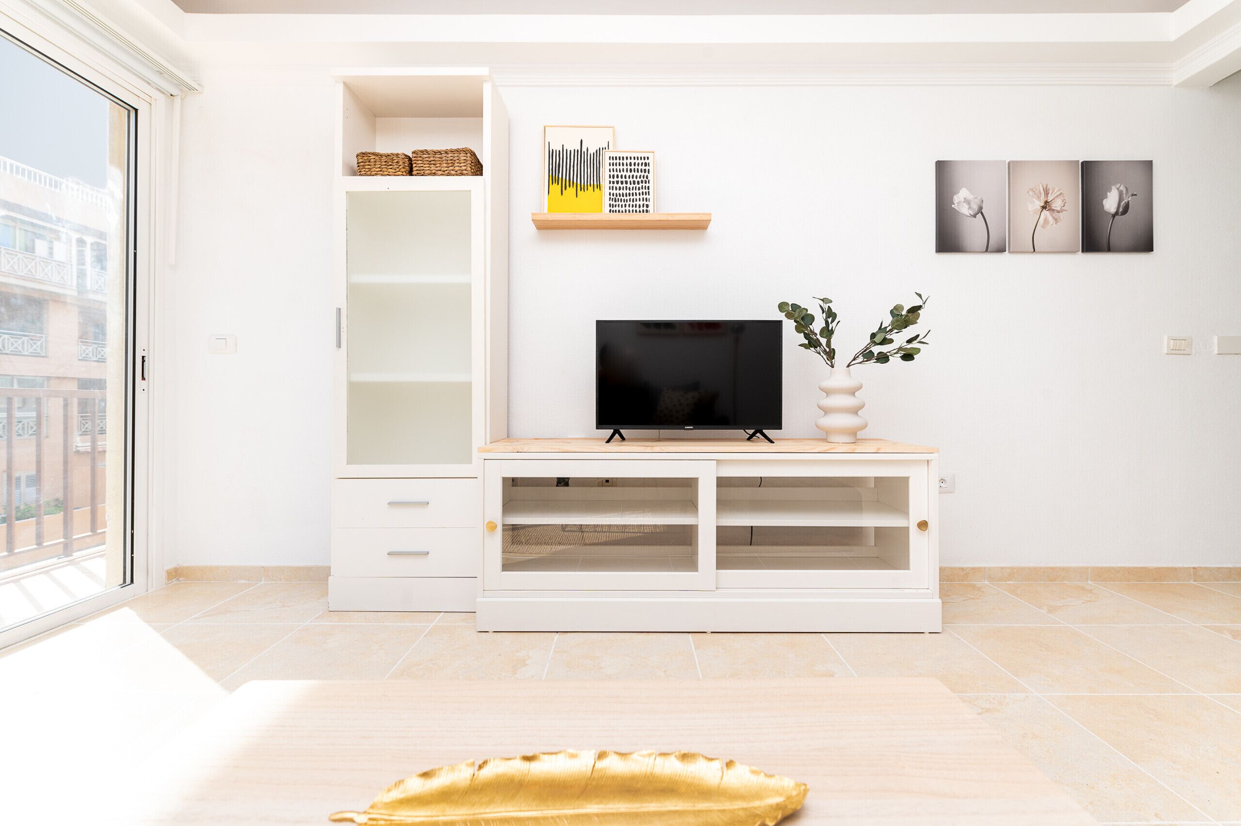 A modern living room with a TV on a wooden stand, a white cabinet with shelves, and a large window.