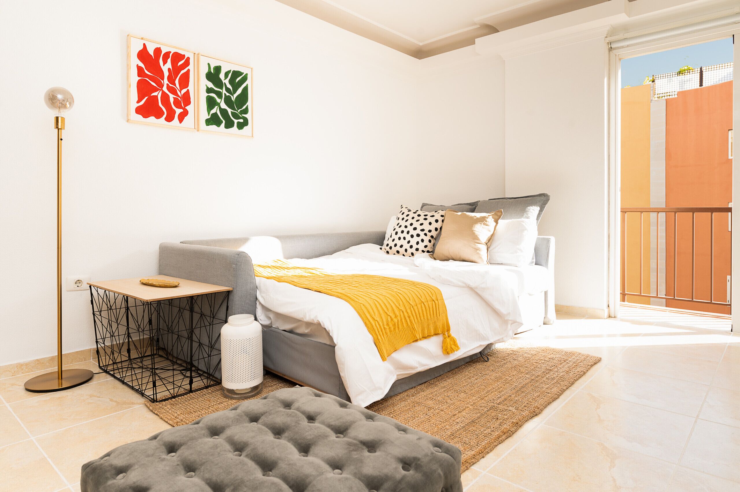 A bedroom with a bed, a grey sofa, a small table, a floor lamp, and artwork on the wall.