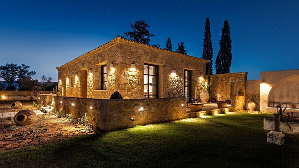 Uma casa de pedra com exterior bem iluminado, um gramado e um céu noturno estrelado.