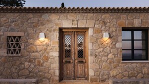 Um edifício de pedra com uma porta dupla de madeira, duas luzes fixadas na parede e uma janela com padrão de grade.