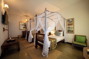 Un lit à baldaquin, une chaise recouverte d’un tissu vert, une table de chevet en bois et un téléviseur fixé au mur.
