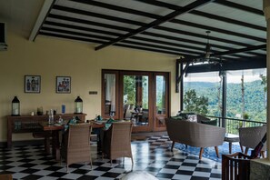 A dining area with checkered flooring, wicker furniture, and a view of a mountainous landscape through large windows. - Riverdale Bungalow (Kandy)