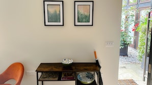 Um quarto com uma mesa de madeira escura, duas obras de arte emolduradas, uma cadeira e vista para um jardim através de uma porta de vidro.
