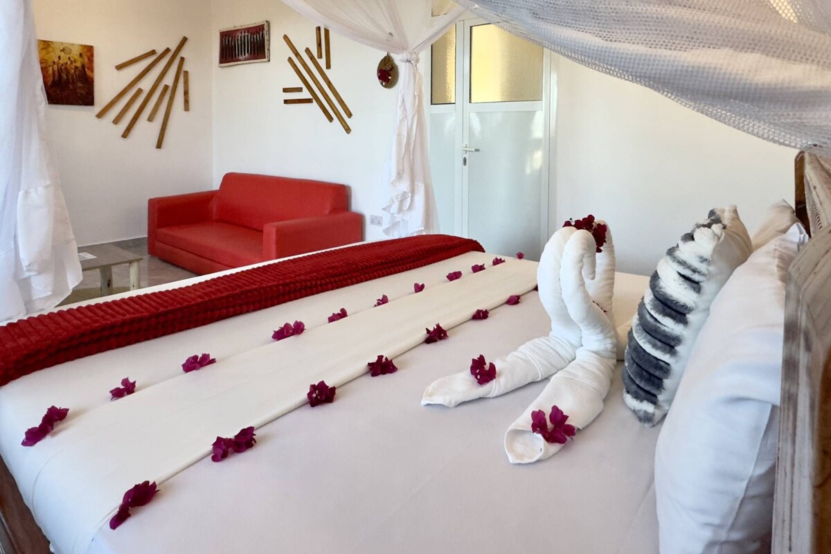 Un letto rifatto con cura, una coperta rossa e petali di rosa, un divano rosso e un letto a baldacchino bianco.