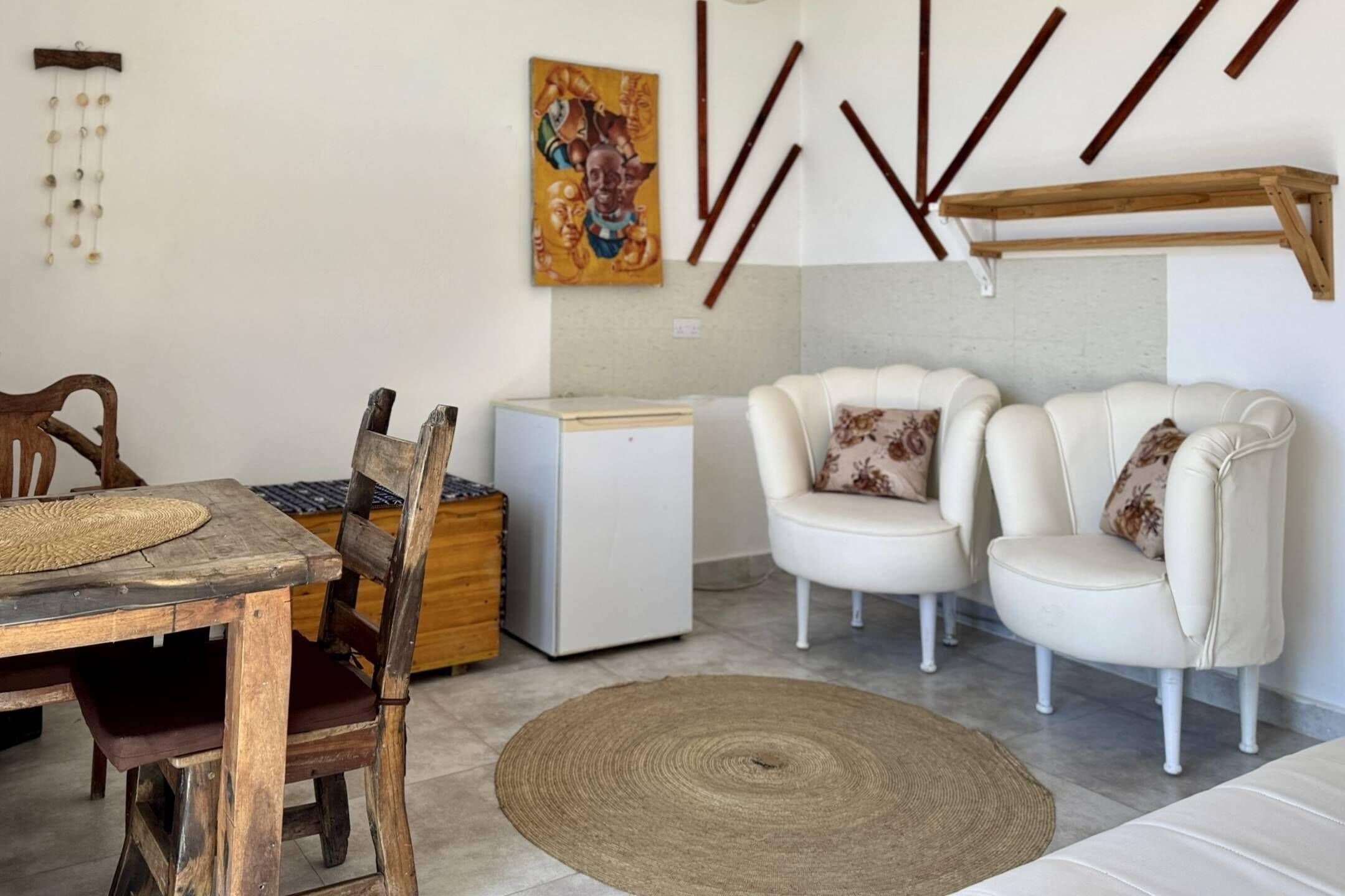 A living room with a wooden dining table, two white armchairs, a white sofa, a small white refrigerator, and a woven rug.