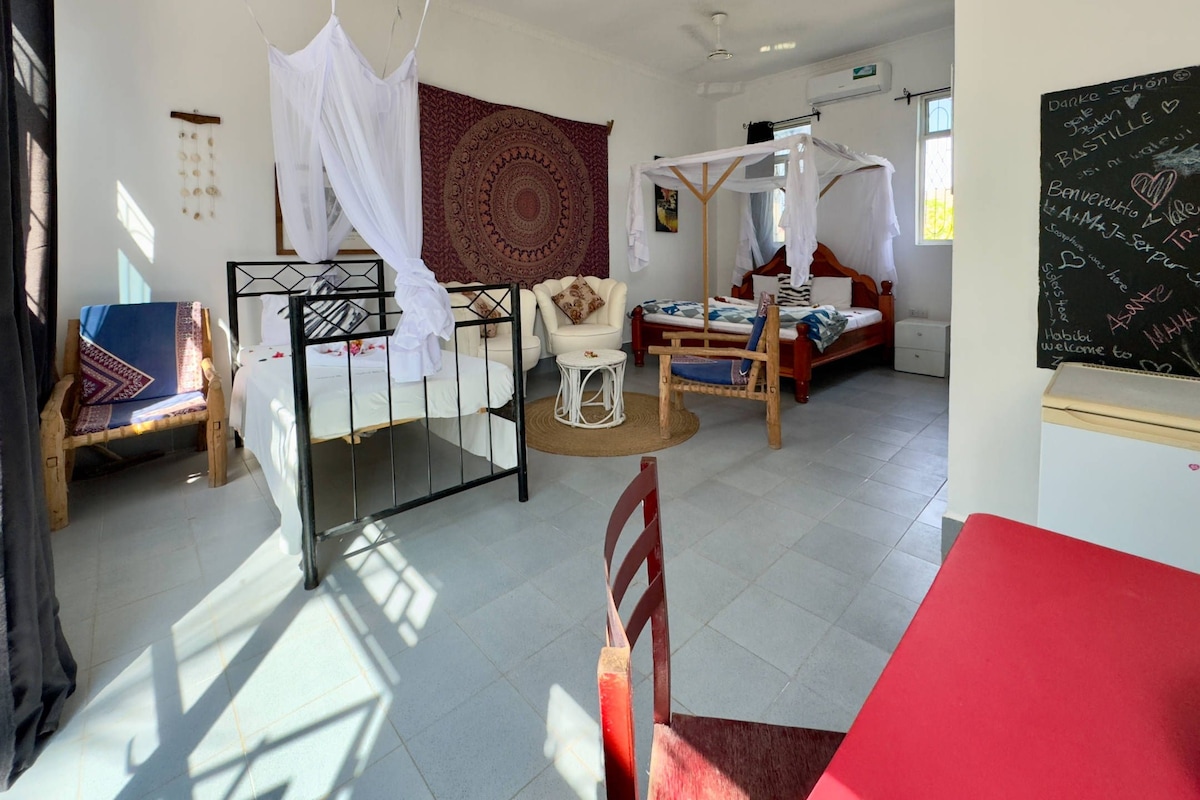 A room with a bed, a chair, a small table, a red table, and a tapestry on the wall.