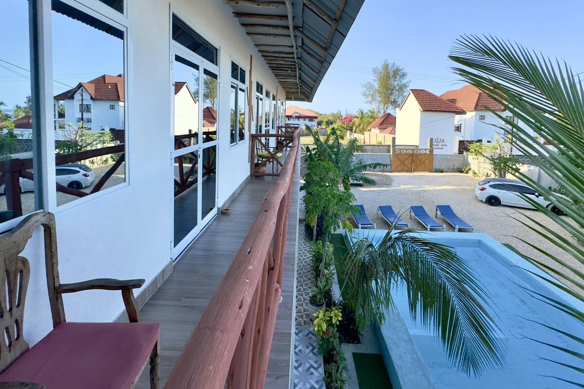A hotel with a covered balcony, outdoor seating, a swimming pool, and a view of neighboring houses.