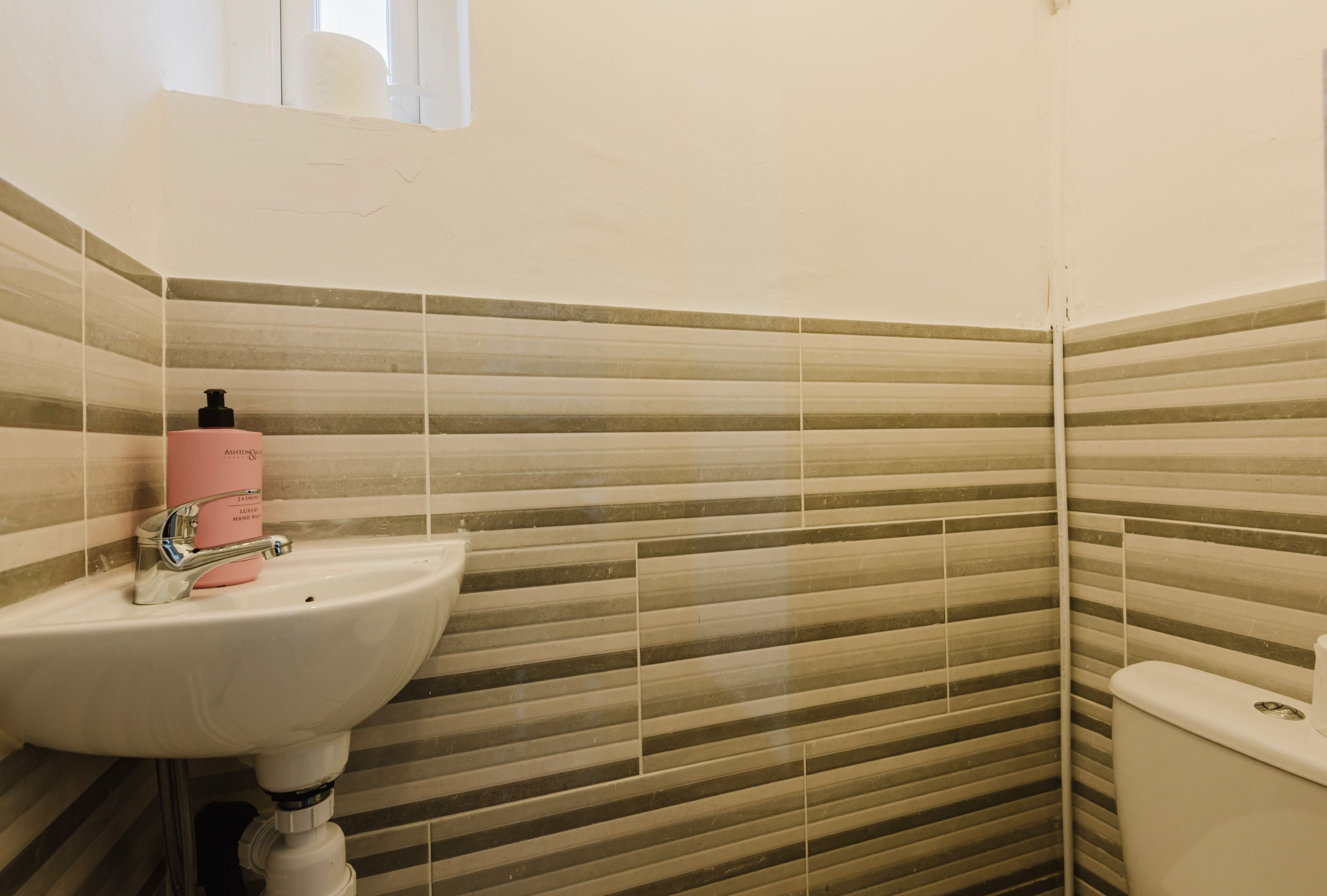 Un baño con lavamanos blanco, una botella rosa sobre el lavamanos y paredes con azulejos a rayas.