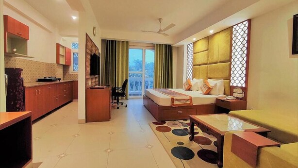 A modern hotel room with a bed, a sofa, a dining table, a kitchen area, and a balcony with curtains. - The Angel Hotel And Suites (Gurugram)