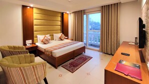 A hotel room with a bed, a desk, a chair, a ceiling fan, and a view of the outside. - The Angel Hotel And Suites (Gurugram)
