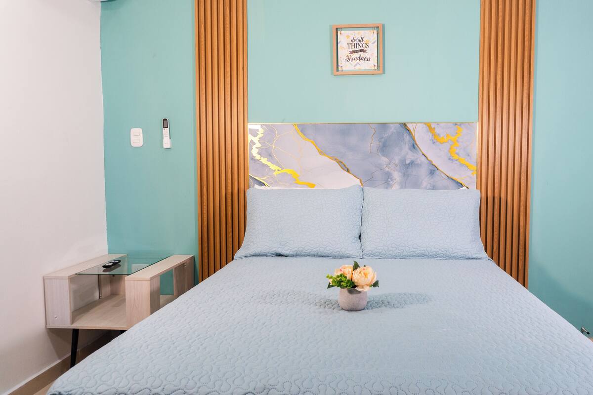 A bedroom with a bed, a bedside table, a nightstand, a wall-mounted picture, and a wooden headboard.