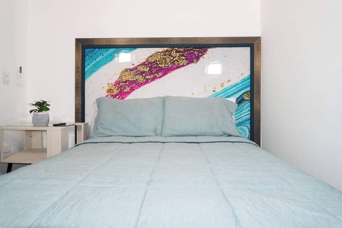 A bed with a light blue bedspread and two pillows, a bedside table with a plant, and a wall with a colorful abstract painting.