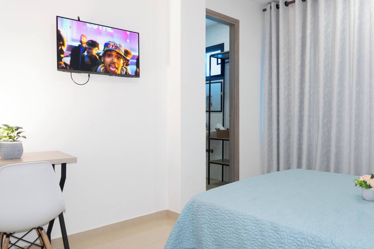 A hotel room with a bed, a desk, a chair, a TV, and a bathroom visible through an open door.