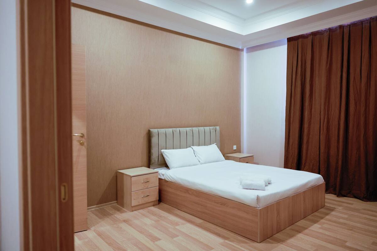 Une chambre moderne avec un lit en bois, des tables de chevet et une grande fenêtre avec des rideaux.