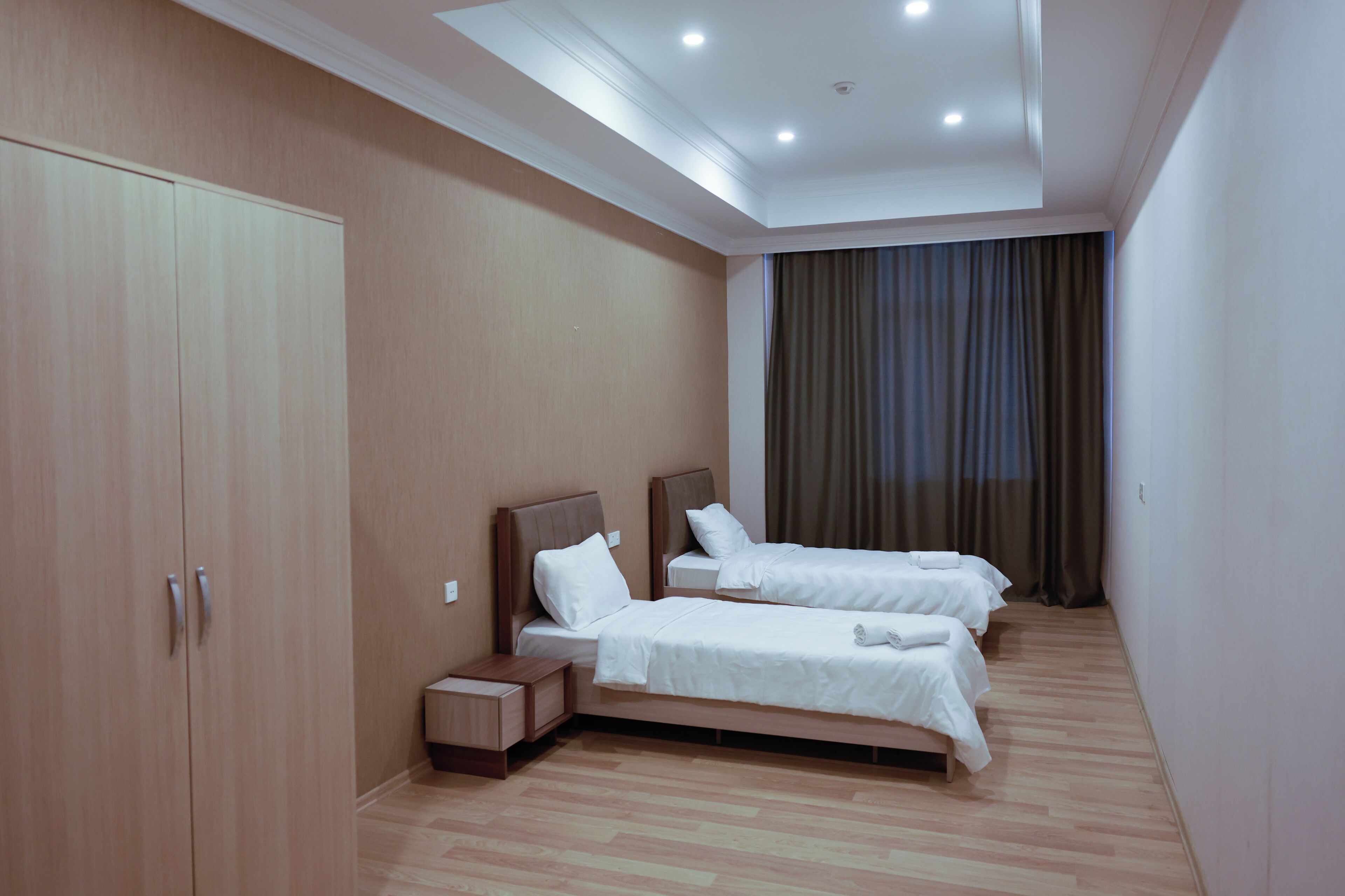 Habitación de hotel con dos camas individuales, un armario, una mesita de noche y un gran ventanal con cortinas.