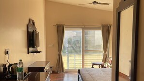 A hotel room with a bed, a desk with a chair, a TV mounted on the wall, a large mirror, and a sliding glass door leading to an outdoor area. - Vrindavan Greens Resort (Udaipur)